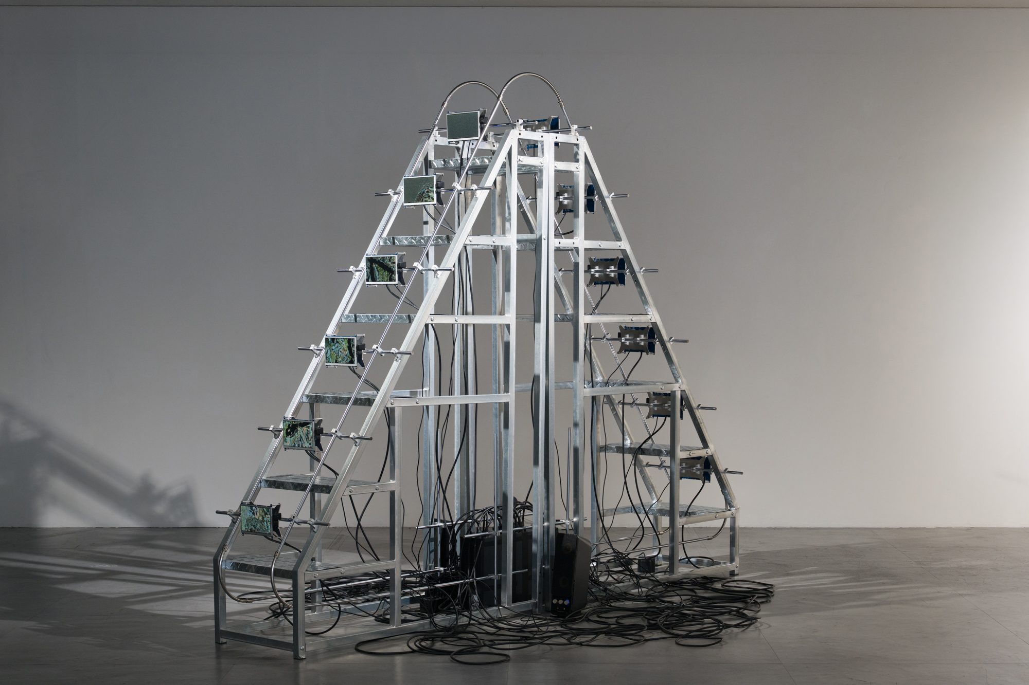 A steel staircase standing alone in a grey room, small screens mounted along both of its sides.