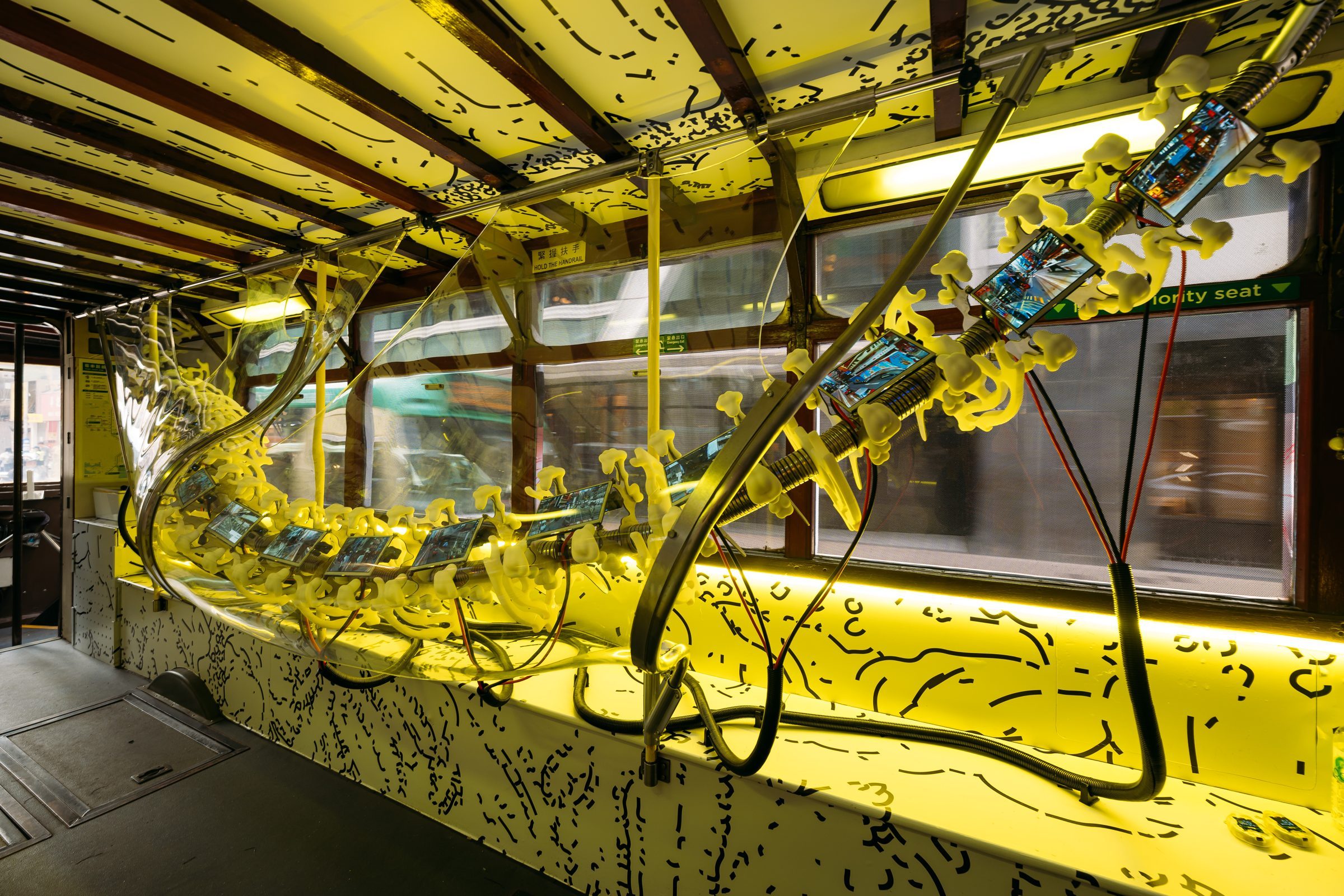 Inside the lower deck of a tram, a long spine-like sculpture lined with small screens runs along the seats under yellow light.