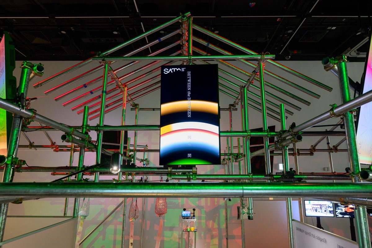 A scaffolding structure lit green and red, with a vertical screen at its centre showing the SATA Fest title, Between the Between.