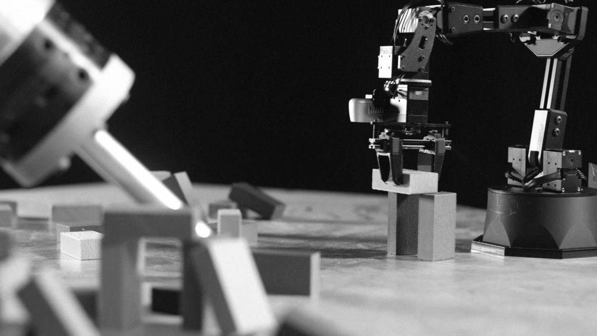 A small robot arm builds a block column while the large tool knocks down an arch in the foreground.