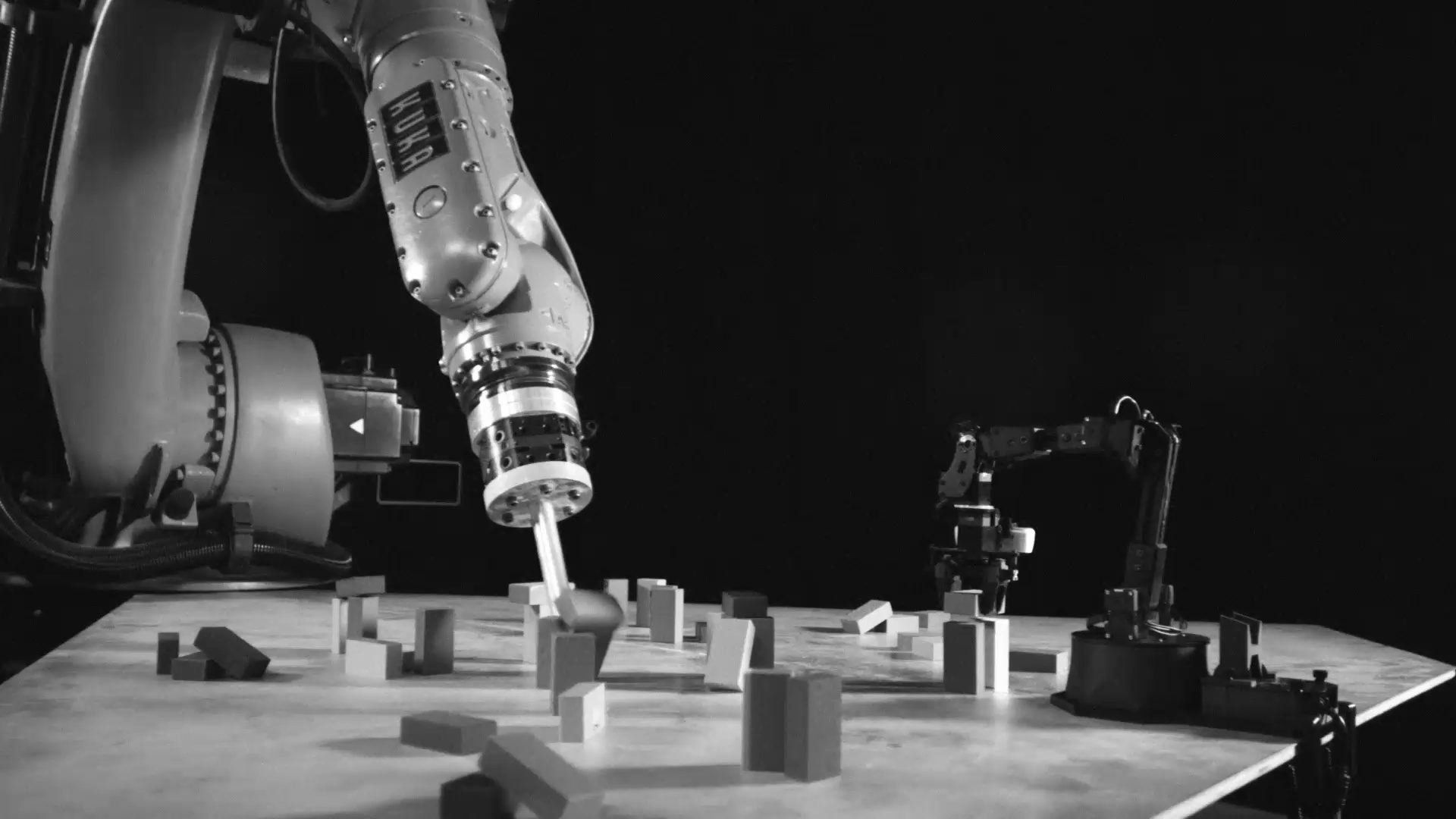 In black and white, an industrial robot arm reaches into scattered wooden blocks while a small robot arm works at the edge of the table.